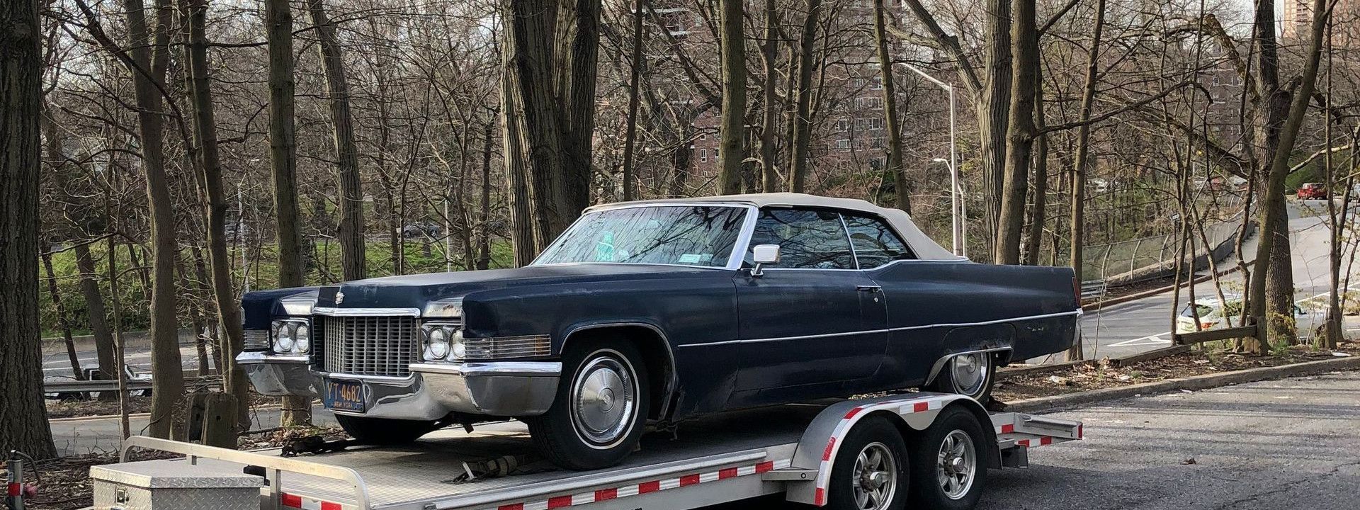 Classic blue car on a trailer amidst leafless trees on a street.